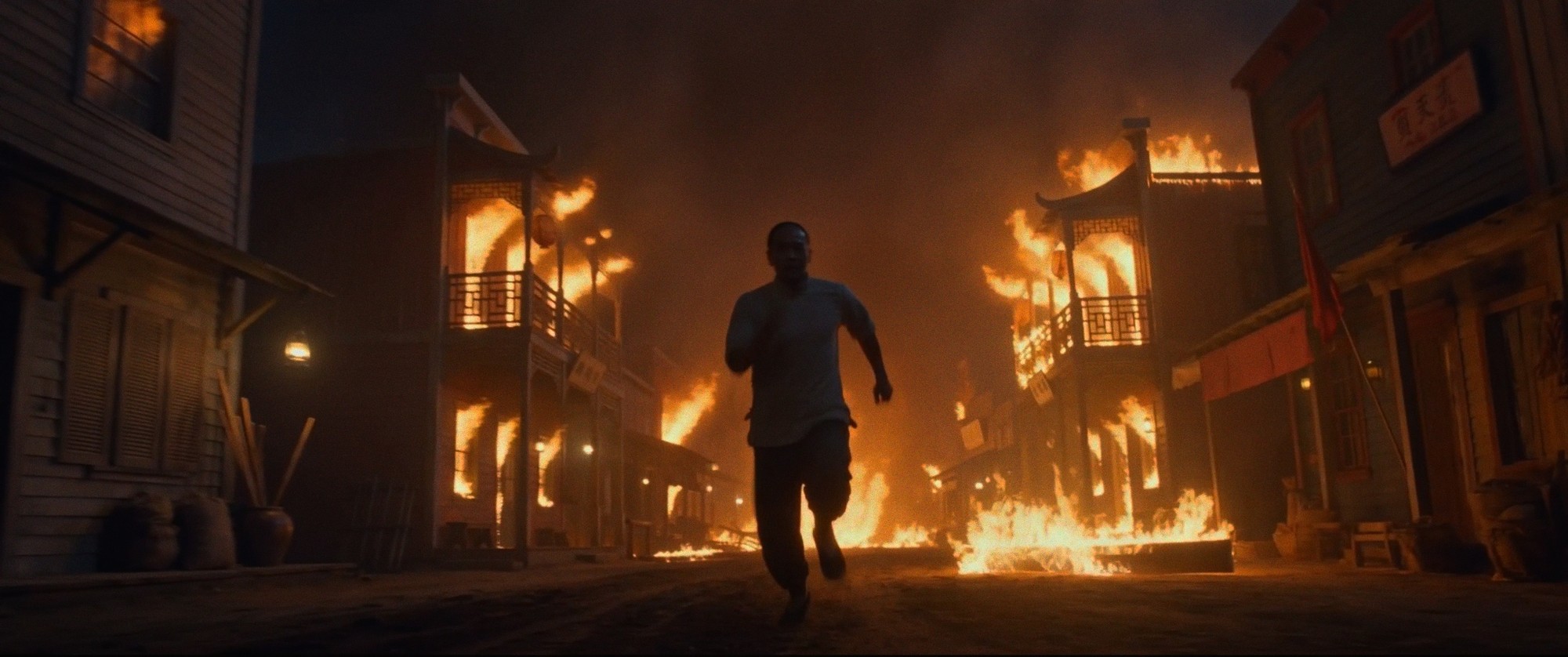 A man runs down a burning street of wooden buildings at night, flames on both sides.